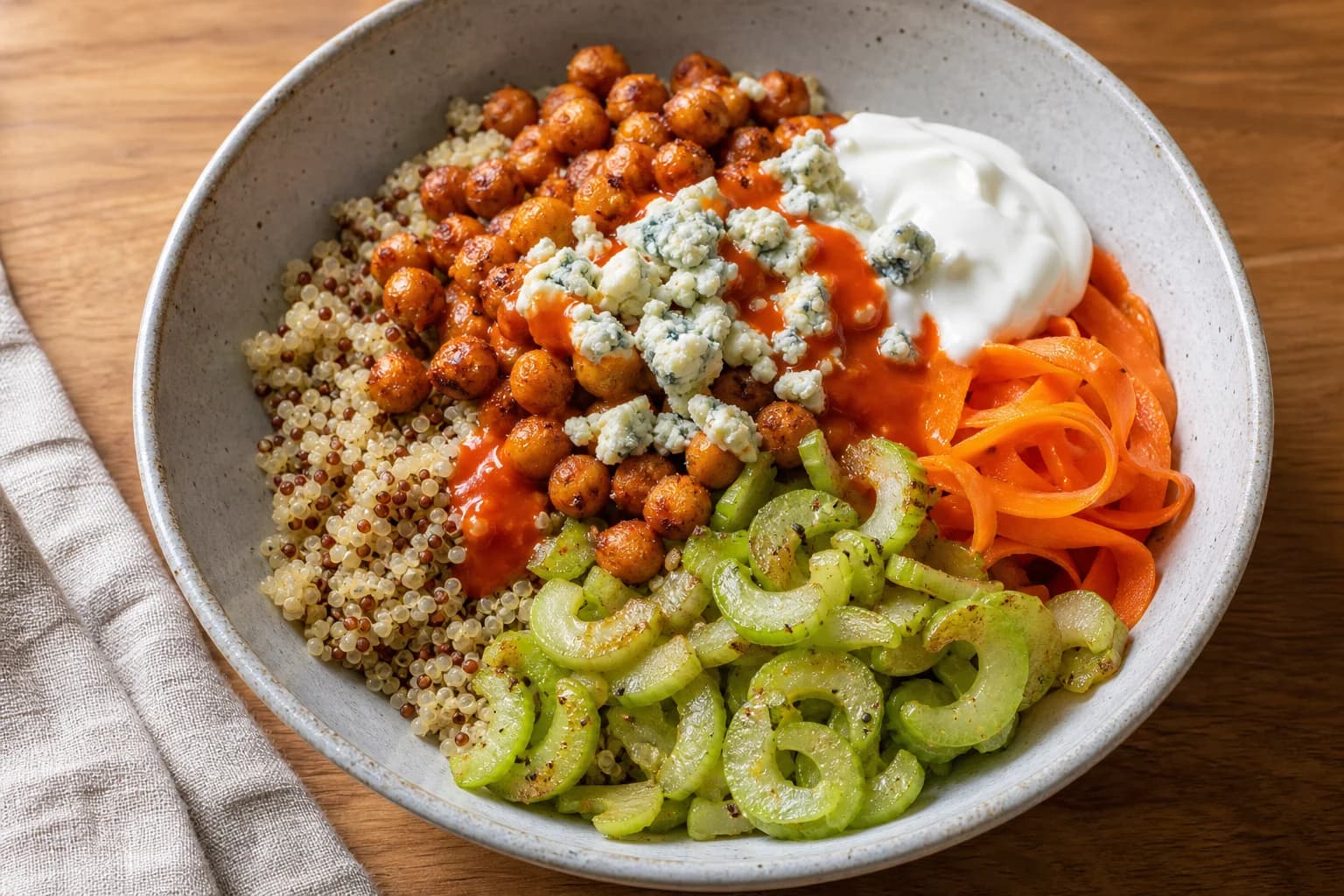 Single bowl of buffalo chickpea quinoa: quinoa base with roasted chickpeas in buffalo sauce, sautéed celery slices, shaved carrot ribbons, a dollop of Greek yogurt and crumbled blue cheese on a wooden surface.